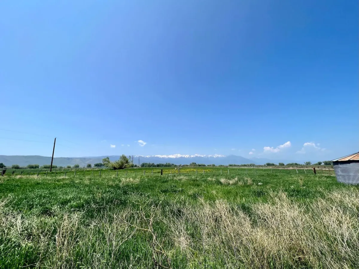 A panoramic view of distant mountains beneath a broad blue sky, with rolling green fields in the foreground. The open landscape evokes a sense of freedom and possibility, perfect for hiking, biking, and exploring nature.