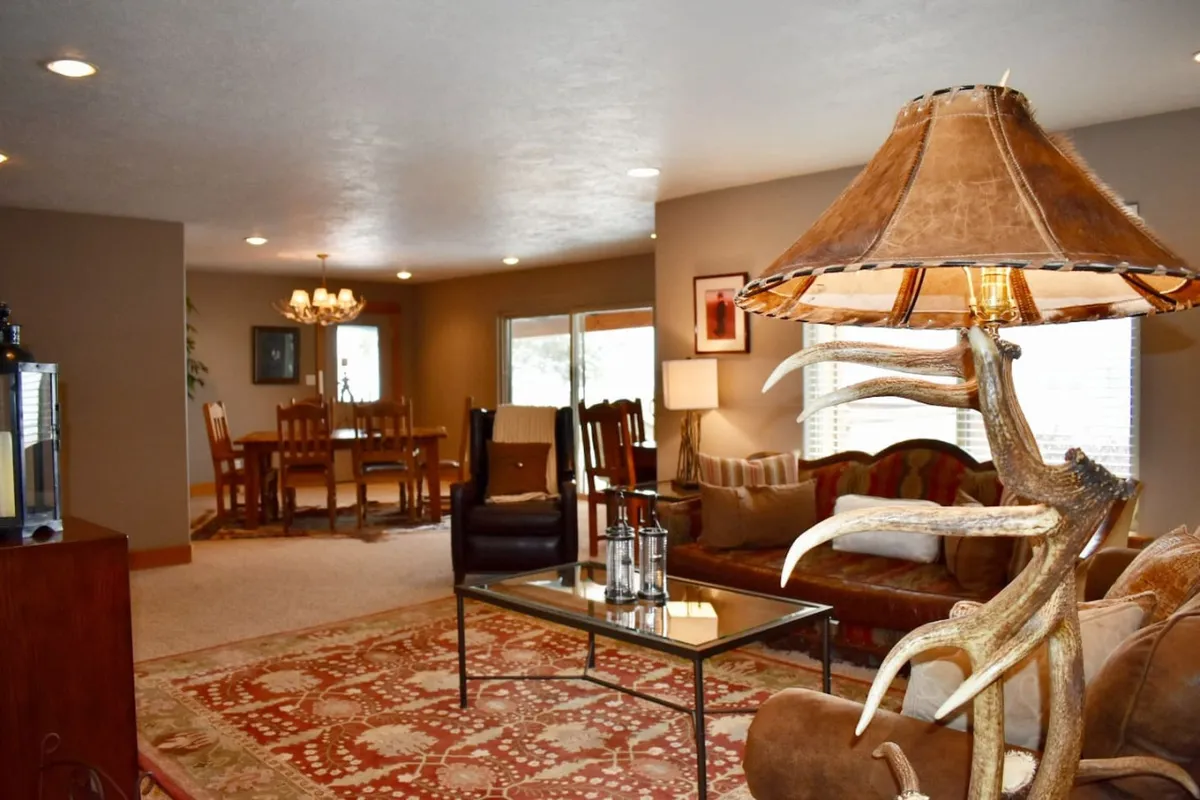 The image captures a warm, inviting living and dining space adorned with rustic country décor. A cozy sofa sits beside a unique antler lamp, while the dining area is set for gatherings, with hints of a nearby fireplace and spacious kitchen just out of view.