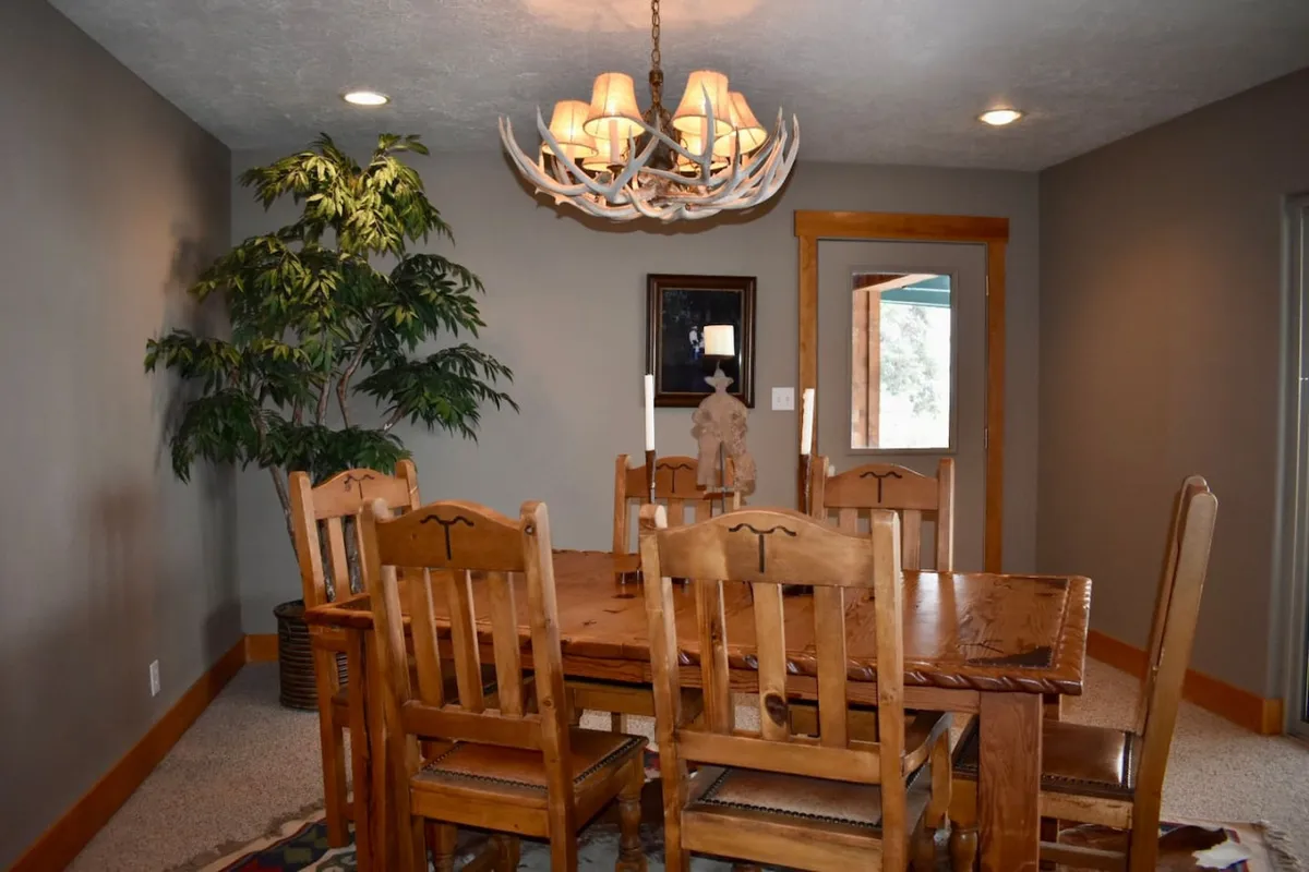 A warm dining area centered around a rustic wooden dining table, illuminated by an antler chandelier and decorated with a potted plant. The space radiates a welcoming, communal atmosphere ideal for family gatherings or special meals.