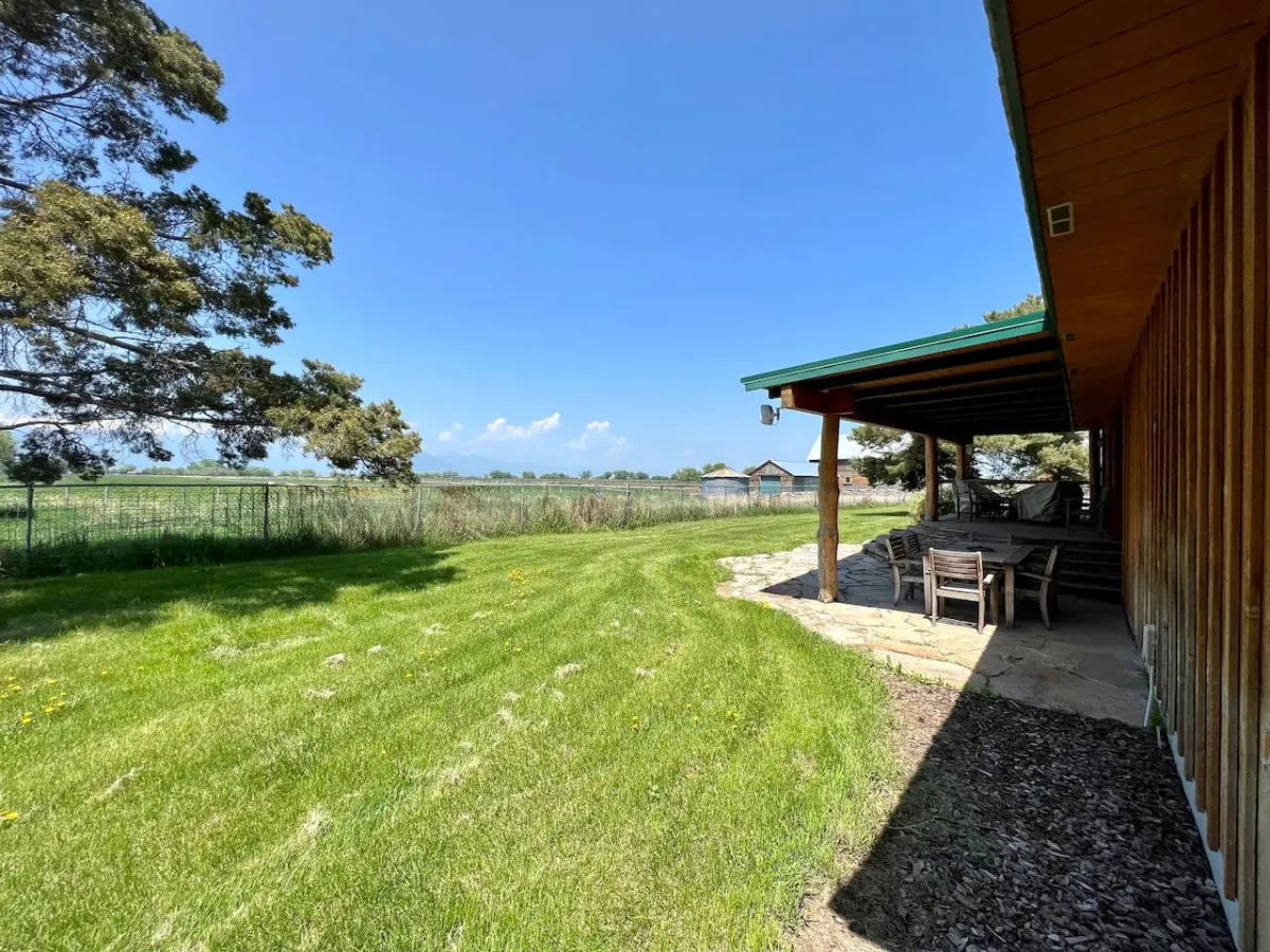 A wooden deck with comfortable outdoor seating overlooks a lush green yard and distant barns. The scene is set for outdoor dining, relaxing in nature, and gathering with friends or family under open skies, with the tranquil ranch landscape as a backdrop.