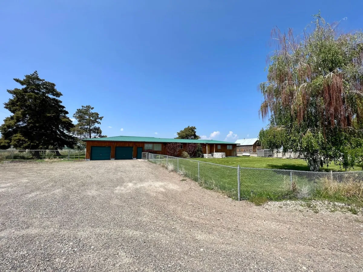This image shows the house with a green roof, a fenced yard, spacious lawn, large trees, and a gravel driveway. The scene feels open and inviting, with plenty of space for kids and pets to play, as well as room for family gatherings or outdoor barbecues.