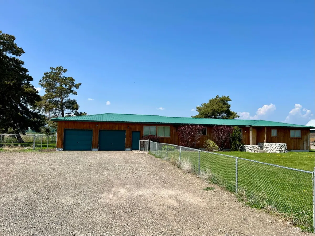 An exterior shot showing the wooden house with a green metal roof, two garage doors, landscaped trees, and a fenced yard. The fenced outdoor area is prominent, signaling a secure space for pets and family play.