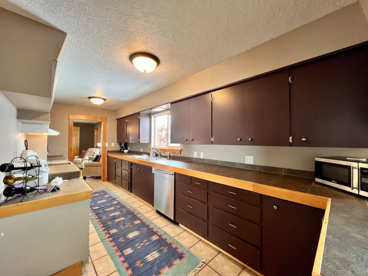 A bright, welcoming kitchen featuring a central island with a sink, modern appliances, and a window offering natural light. The space is designed for group cooking and gatherings, with ample counter space and accessible amenities.