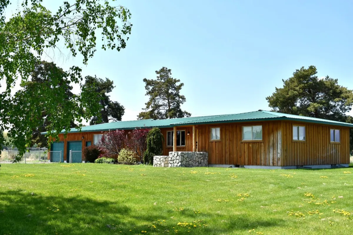 A charming wooden exterior with a green roof, lush grass, and mature trees creating a welcoming and homey environment. The surrounding lawn is spacious, providing ample space for outdoor activities, picnics, or family gatherings. The cozy, country style of the home and natural landscaping evoke comfort and relaxation.