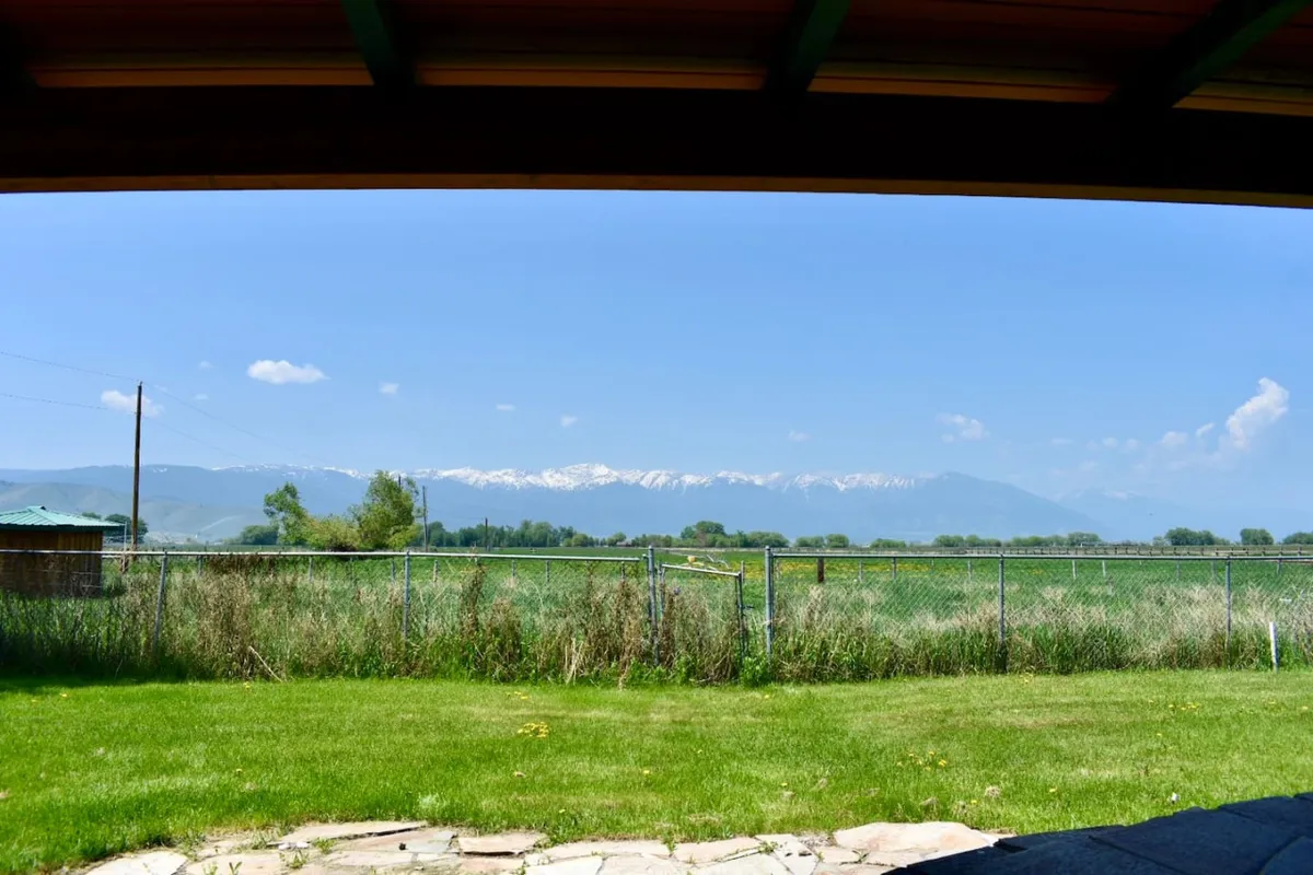 A picturesque view of snow-capped mountains rising above lush green grass, bordered by a classic fence and set against a clear blue sky. The peaceful surroundings capture the natural beauty and wide-open scenery enjoyed at the ranch.