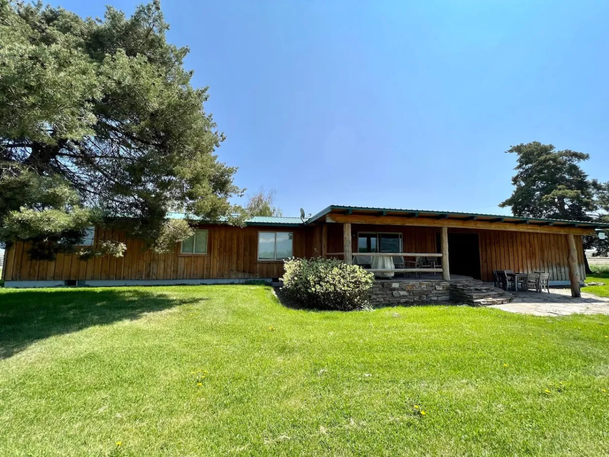 A wide view of the ranch house’s wooden exterior, complete with a green lawn, front porch seating, and trees in the background. The porch is arranged for relaxation, socializing, or simply soaking in the serene landscape as day turns to night.