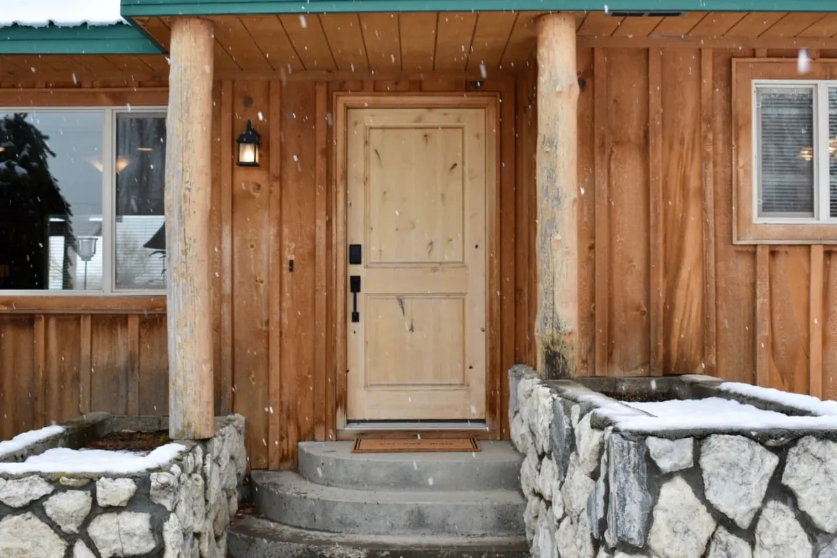 A cozy cabin entrance with a welcoming wooden door, sturdy log pillars, and snow-covered steps, set against a backdrop of wintry landscape. The scene captures both the warmth of the ranch and the excitement of winter activities, such as skiing at Anthony Lakes Ski Area.