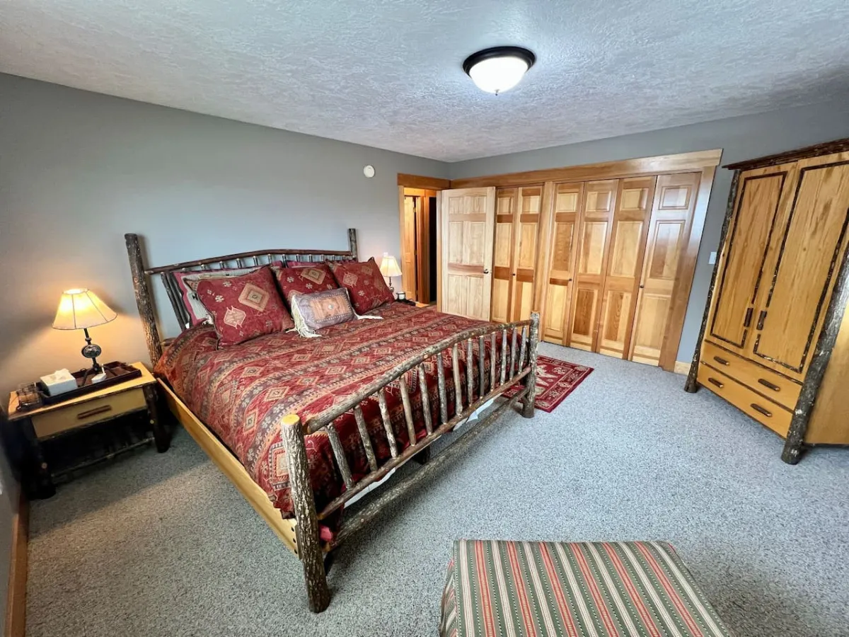 A warm, inviting bedroom featuring a rustic wooden bed frame, decorative pillows, a nightstand with a lamp, and a wardrobe with a wooden finish. The soft bedding and wood accents create a cozy ranch ambiance, highlighting comfort and relaxation.