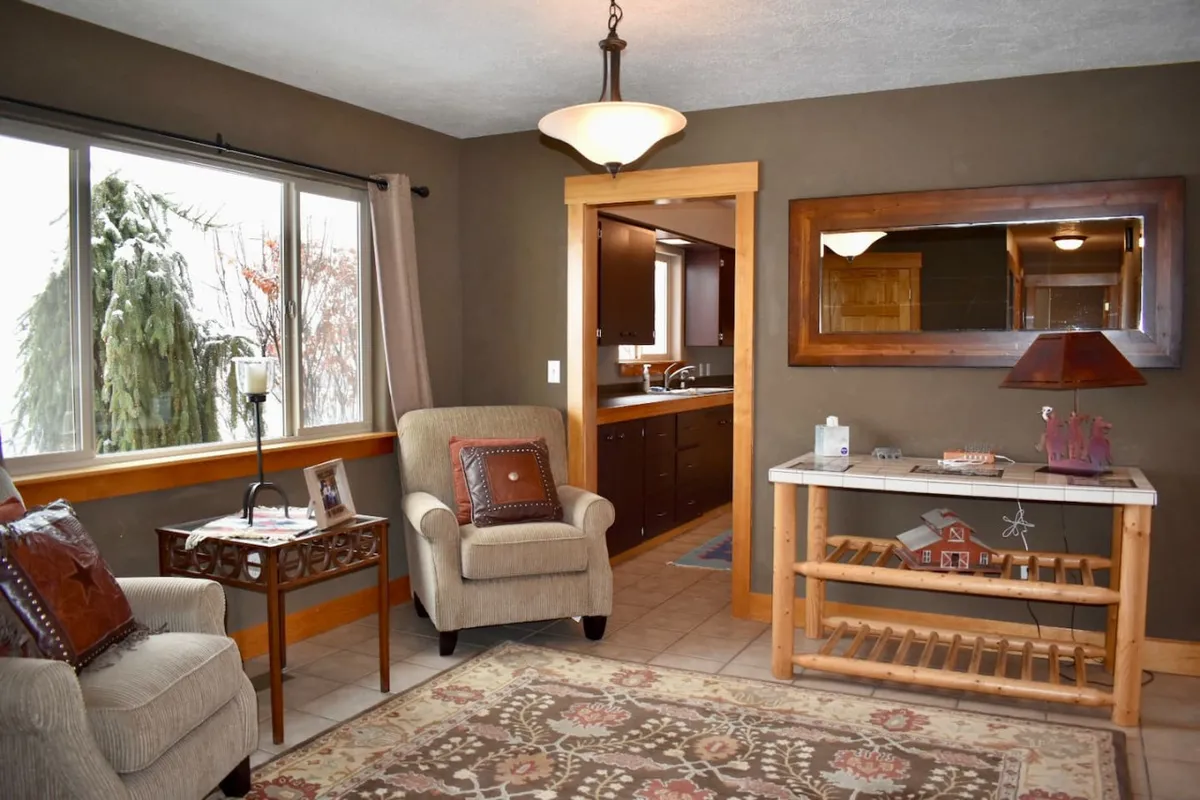 A welcoming main sitting area with plush armchairs gathered around a wooden table, a decorative mirror reflecting the rustic ambiance, and a view through the doorway into the kitchen. The furnishings and decor evoke comfort and rural charm, inviting visitors to relax and socialize.