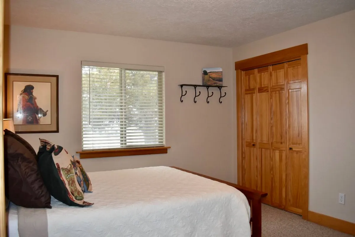 A tranquil bedroom with a comfortable bed, window letting in natural light, framed artwork, and a wooden closet. The mood is peaceful and serene, emphasizing the space and calmness ideal for recharging.